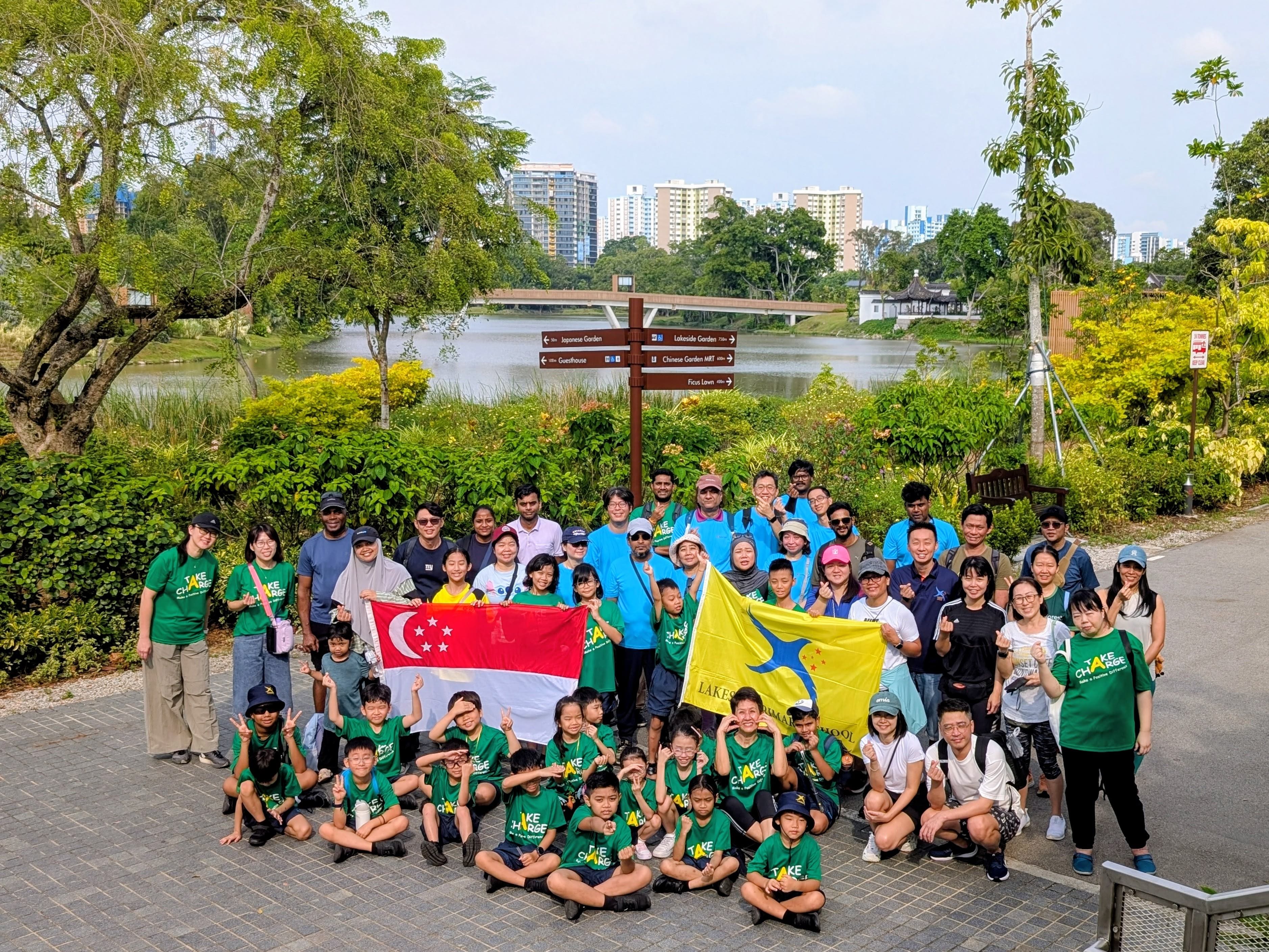 Group photo of our school community and PacificLight representatives posing outdoors near a lake, holding the Singapore and Lakeside Primary School flags